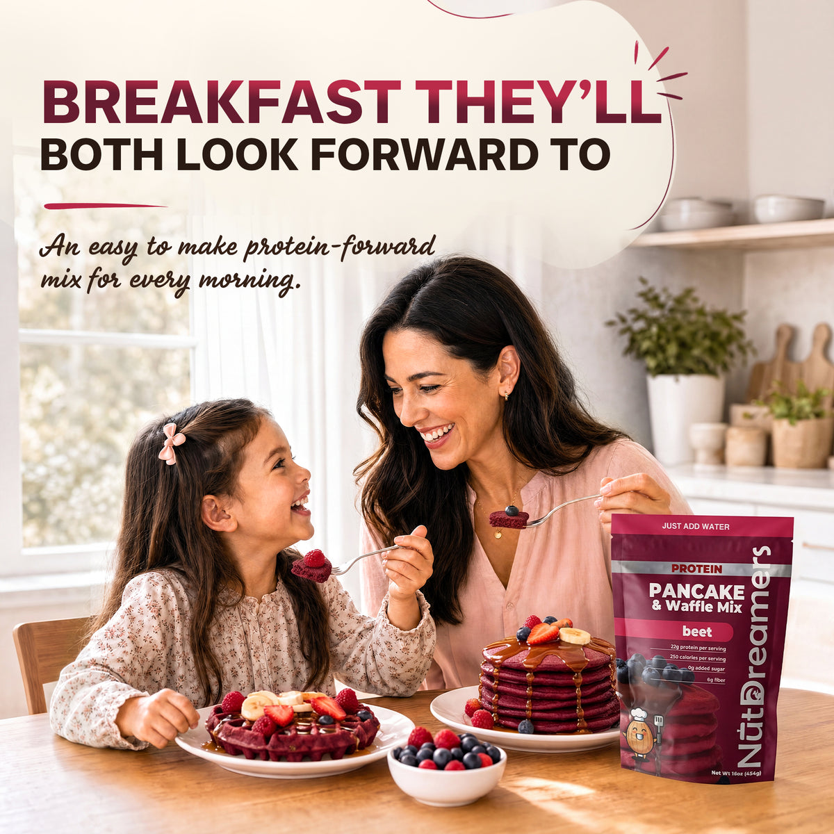 A woman and a young girl sit at a wooden table in a bright kitchen, smiling and feeding each other pancakes. Large promotional text at the top reads 'BREAKFAST THEY'LL BOTH LOOK FORWARD TO,' and a bag of NutDreamers Pancake & Waffle Mix is shown on the table next to pancakes and fruit.
