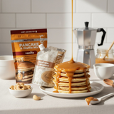 Stack of pancakes with  Nut Dreamers Cashew Caramel, Vanilla Protein pancake and waffle mix packages, and coffee on a kitchen counter.